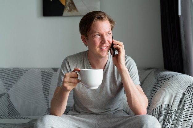 Hombre guapo joven feliz sosteniendo café y hablando por teléfono en casa