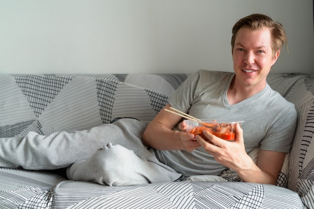 Hombre guapo joven feliz sosteniendo kimchi mientras está acostado en el sofá en casa