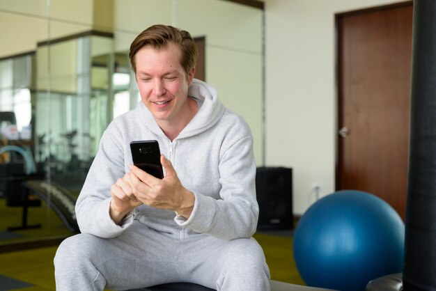 Hombre guapo joven feliz con sudadera con capucha usando teléfono y sentado en el gimnasio