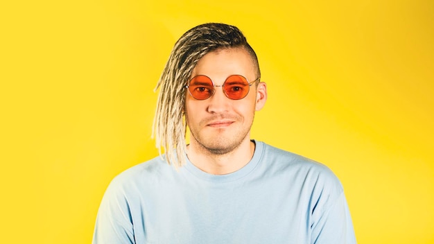 Hombre guapo joven en gafas de sol naranjas mirando a la cámara sonriendo sobre fondo amarillo Hombre alegre con cerdas de rastas en camiseta azul posando en el estudio