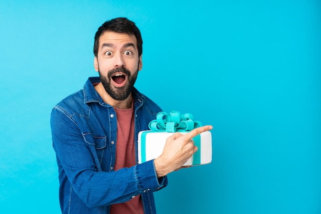 Hombre guapo joven con un gran pastel sobre pared azul aislado sorprendido y apuntando hacia el lado