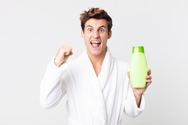 Hombre guapo joven gritando agresivamente con una expresión de enojo con albornoz y sosteniendo una botella de champú