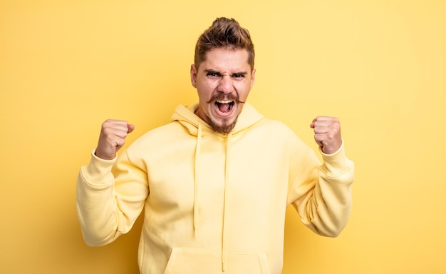 Hombre guapo joven gritando agresivamente con una expresión de enojo o con los puños cerrados celebrando el éxito concepto de bigote extraño