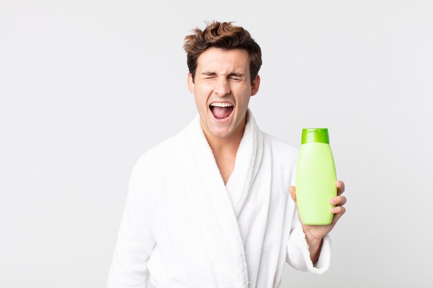 Hombre guapo joven gritando agresivamente, luciendo muy enojado con albornoz y sosteniendo una botella de champú