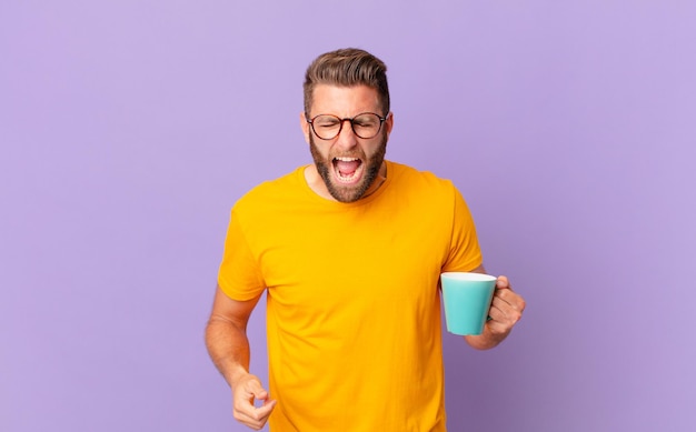 Hombre guapo joven gritando agresivamente, luciendo muy enojado. y sosteniendo una taza de café