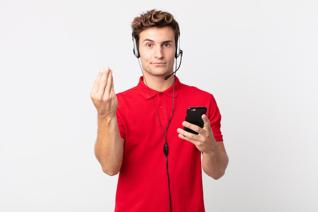 Hombre guapo joven haciendo gesto de capice o dinero, diciéndole que pague con un teléfono inteligente y auriculares