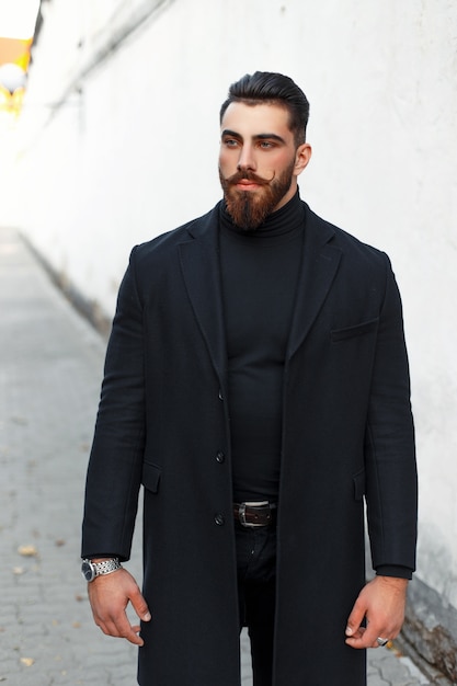 Hombre guapo joven inconformista con cabello y barba en ropa de moda negra con abrigo en la calle