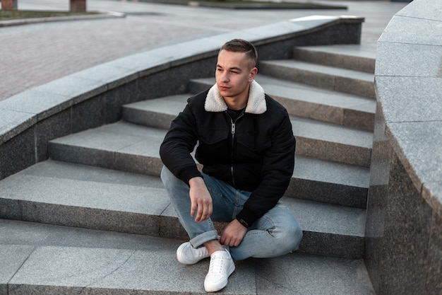 Hombre guapo joven en jeans azul con una chaqueta negra en zapatillas blancas con un peinado sentado en una escalera de piedra vintage en la ciudad