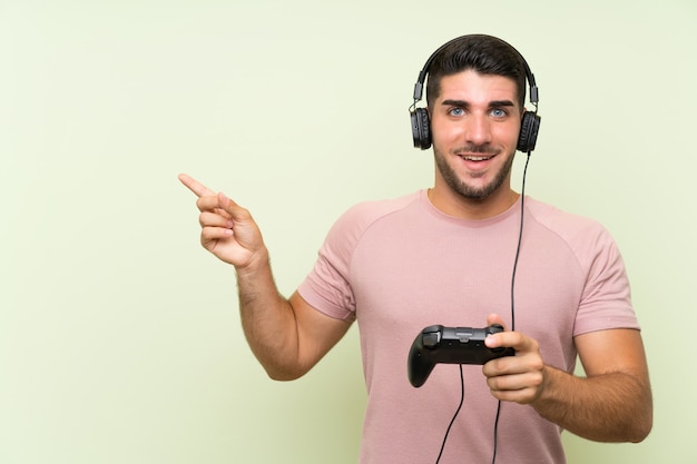 Hombre guapo joven jugando con un controlador de videojuego sobre pared verde aislado sorprendido y apuntando con el dedo hacia un lado