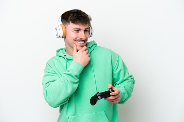 Hombre guapo joven jugando con un controlador de videojuegos aislado sobre fondo blanco mirando hacia un lado y sonriendo