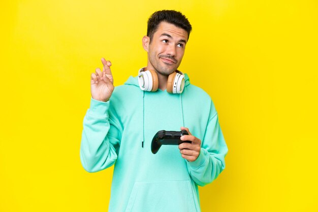 Hombre guapo joven jugando con un controlador de videojuegos sobre una pared aislada con los dedos cruzados y deseando lo mejor