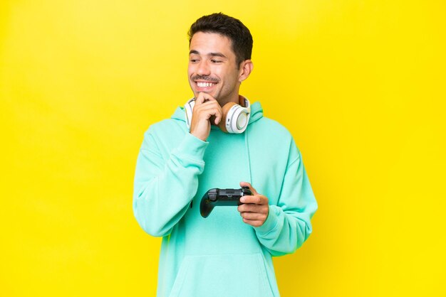 Hombre guapo joven jugando con un controlador de videojuegos sobre una pared aislada mirando hacia un lado y sonriendo