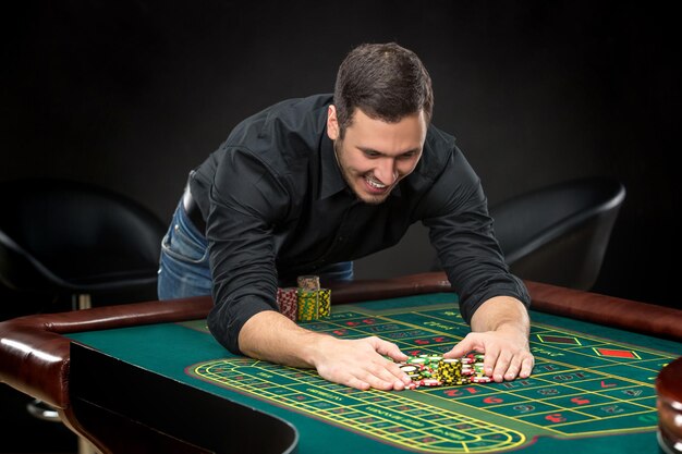 Hombre guapo joven jugando a la ruleta gana en el casino, fichas de juego tomadas por sus manos