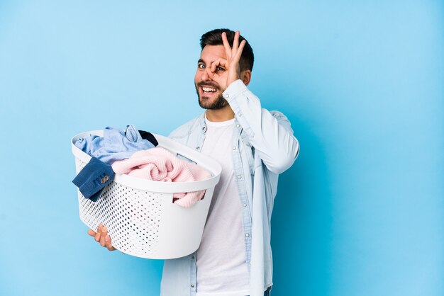 Hombre guapo joven lavando ropa aislado emocionado manteniendo el gesto ok en el ojo.