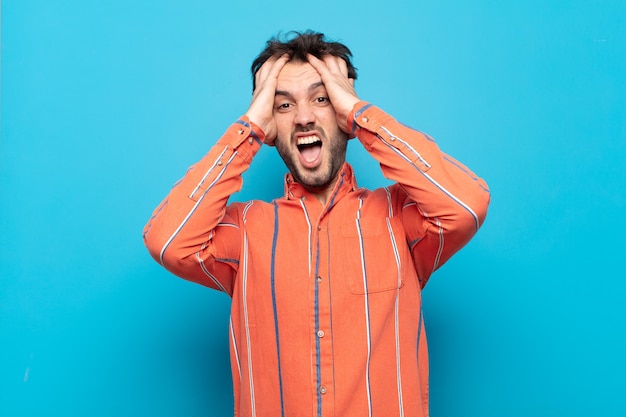 Hombre guapo joven levantando las manos a la cabeza, con la boca abierta, sintiéndose extremadamente afortunado, sorprendido, emocionado y feliz