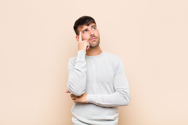 Hombre guapo joven con una mirada concentrada, preguntándose con una expresión dudosa, mirando hacia arriba y hacia el lado contra la pared cálida