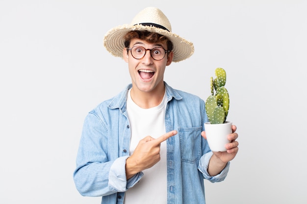 Hombre guapo joven mirando emocionado y sorprendido apuntando hacia un lado. granjero sosteniendo un cactus decorativo