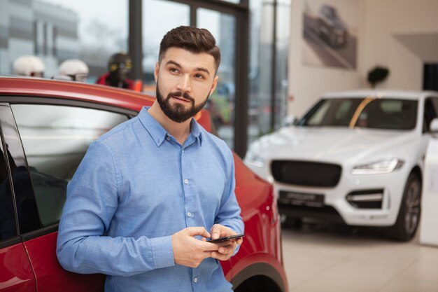 Hombre guapo joven mirando a otro lado pensativamente mientras usa el teléfono inteligente en el concesionario de automóviles, espacio de copia