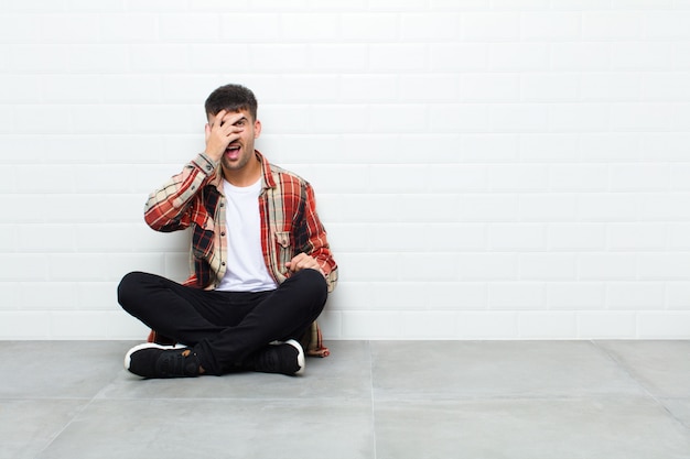 Hombre guapo joven mirando sorprendido, asustado o aterrorizado, cubriéndose la cara con la mano y mirando entre los dedos sentado en el piso de cemento