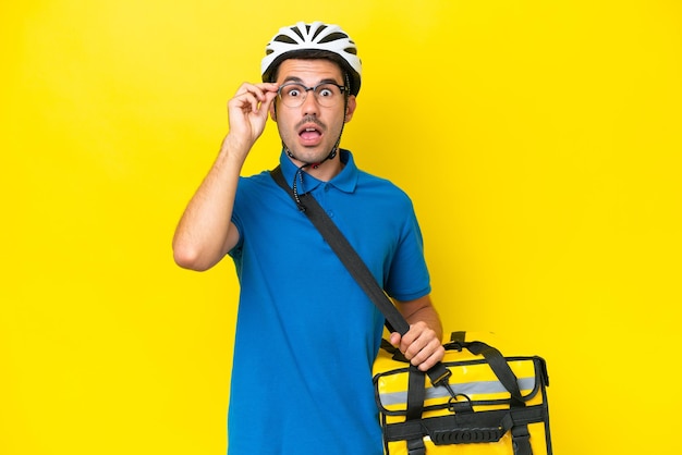 Hombre guapo joven con mochila térmica sobre fondo amarillo aislado con gafas y sorprendido