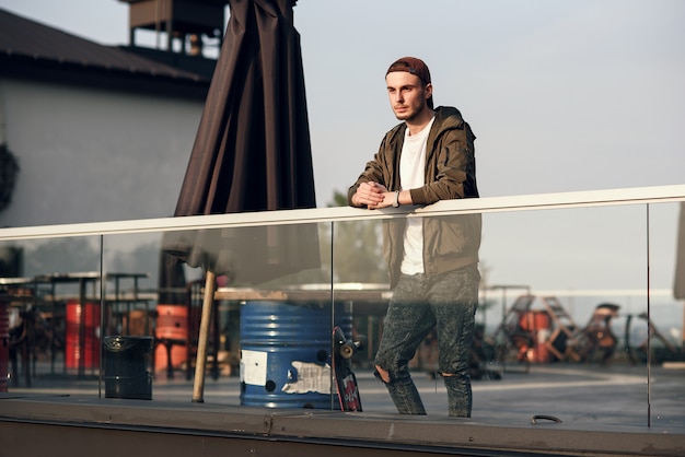 Hombre guapo joven con patín en la azotea del edificio industrial en la puesta del sol.