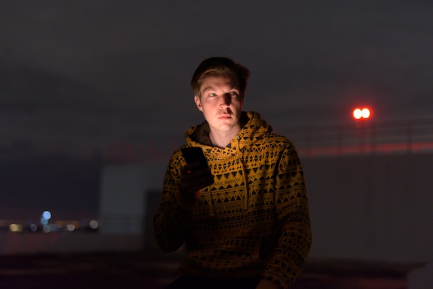 Hombre guapo joven pensando mientras usa el teléfono en la azotea del edificio durante el clima tormentoso en la noche