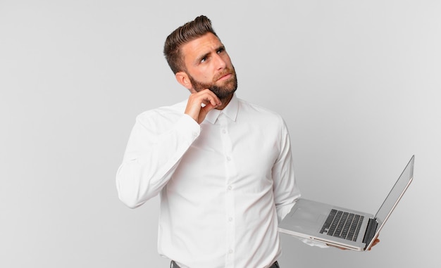 Hombre guapo joven pensando, sintiéndose dudoso y confundido y sosteniendo una computadora portátil