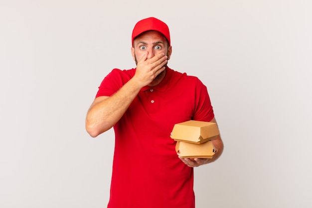 Hombre guapo joven que cubre la boca con las manos con un concepto de entrega de hamburguesa sorprendido