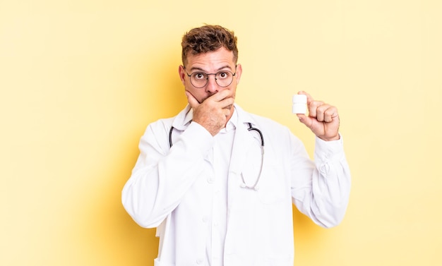 Hombre guapo joven que cubre la boca con las manos con una sorpresa. concepto de botella de píldoras médico