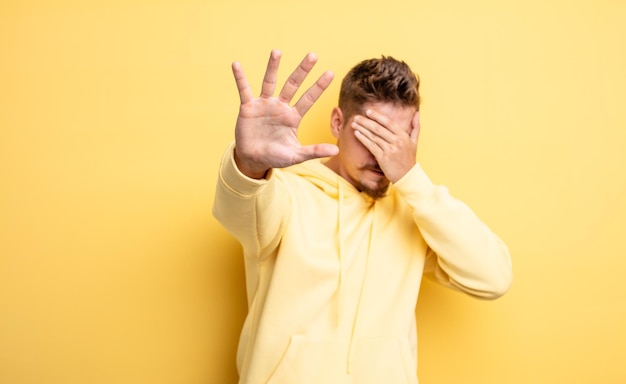 Hombre guapo joven que cubre la cara con la mano y pone la otra mano al frente para detener la cámara, rechazando fotos o imágenes. extraño concepto de bigote