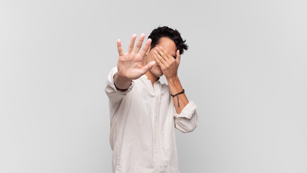 Hombre guapo joven que cubre la cara con la mano y pone la otra mano al frente para detener la cámara, rechazando fotos o imágenes