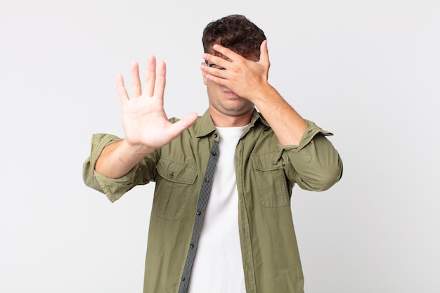 Hombre guapo joven que cubre la cara con la mano y pone la otra mano al frente para detener la cámara, rechazando fotos o imágenes