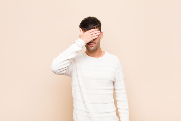 Hombre guapo joven que cubre los ojos con una mano sintiéndose asustado o ansioso, preguntándose o esperando ciegamente una sorpresa sobre la cálida pared