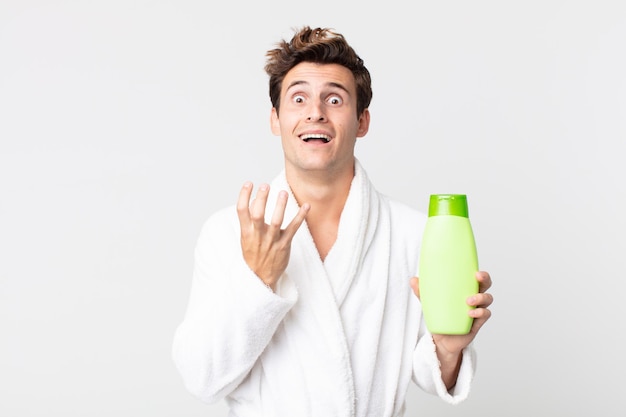 Hombre guapo joven que parece desesperado, frustrado y estresado con albornoz y sosteniendo una botella de champú