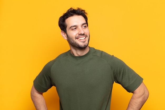 Hombre guapo joven que parece feliz, alegre y seguro, sonriendo con orgullo y mirando al lado con ambas manos en las caderas