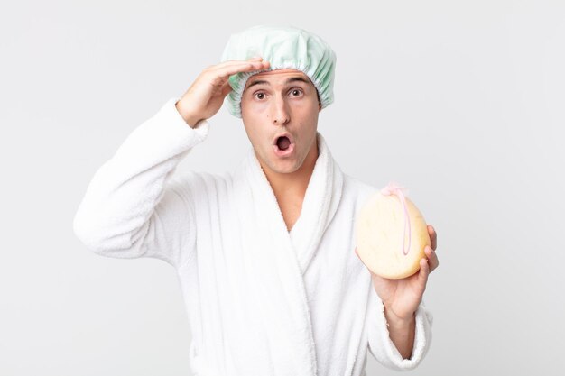 Hombre guapo joven que parece feliz, asombrado y sorprendido con albornoz, gorro de ducha y una esponja