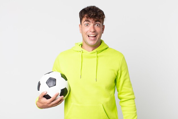 Hombre guapo joven que parece feliz y gratamente sorprendido y sosteniendo un balón de fútbol