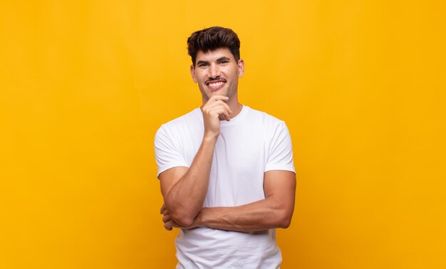 Hombre guapo joven que parece feliz y sonriente con la mano en la barbilla, preguntándose o haciendo una pregunta, comparando opciones