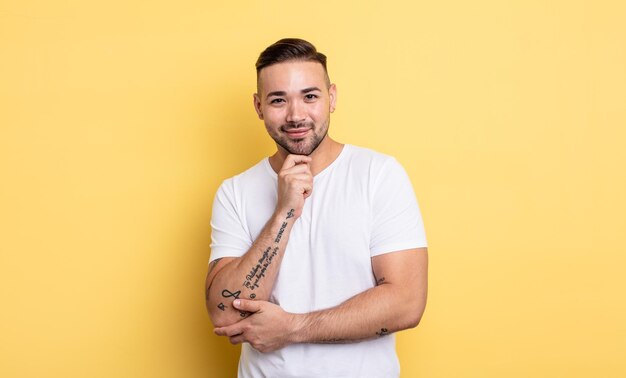 Hombre guapo joven que parece feliz y sonriente con la mano en la barbilla, preguntándose o haciendo una pregunta, comparando opciones