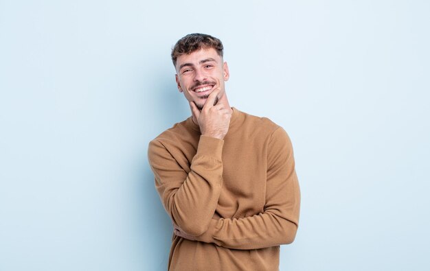 Hombre guapo joven que parece feliz y sonriente con la mano en la barbilla, preguntándose o haciendo una pregunta, comparando opciones