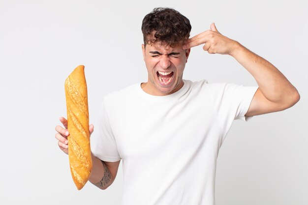 Hombre guapo joven que parece infeliz y estresado, gesto de suicidio haciendo un signo de pistola y sosteniendo una baguette de pan