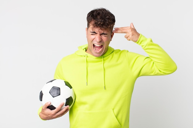 Hombre guapo joven que parece infeliz y estresado, gesto de suicidio haciendo un signo de pistola y sosteniendo un balón de fútbol