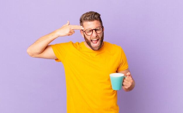 Hombre guapo joven que parece infeliz y estresado, gesto de suicidio haciendo signo de pistola. y sosteniendo una taza de café