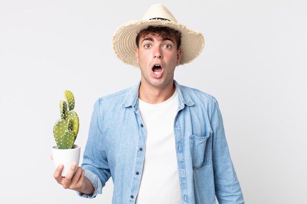 Hombre guapo joven que parece muy conmocionado o sorprendido. granjero sosteniendo un cactus decorativo