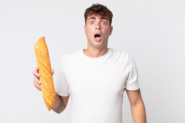 Hombre guapo joven que parece muy conmocionado o sorprendido y sosteniendo una baguette de pan