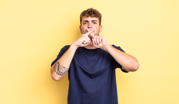 Hombre guapo joven que parece serio y disgustado con ambos dedos cruzados al frente en rechazo, pidiendo silencio