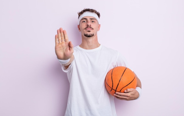 Hombre guapo joven que parece serio mostrando la palma abierta haciendo gesto de parada. concepto de baloncesto