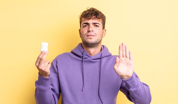 Hombre guapo joven que parece serio mostrando la palma abierta haciendo gesto de parada. concepto de botella de pastillas