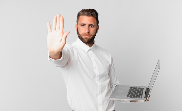 Hombre guapo joven que parece serio mostrando la palma abierta haciendo gesto de parada y sosteniendo una computadora portátil