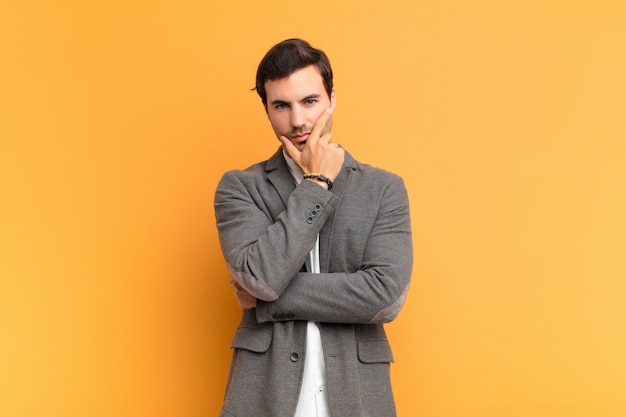 Hombre guapo joven que parece serio, pensativo y desconfiado, con un brazo cruzado y la mano en la barbilla, opciones de ponderación